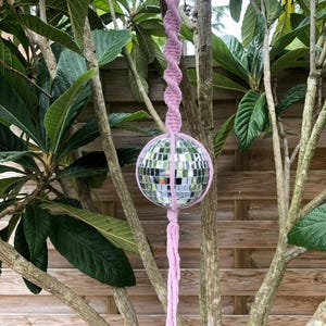 Puede incluir: Bola de discoteca colgante con un cordón y borla de macramé rosa. La bola de espejo está suspendida de un anillo de madera, creando un acento decorativo al aire libre. El macramé es de color rosa claro.
