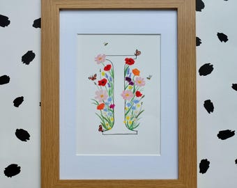 Puede incluir: Una lámina artística enmarcada con la letra mayúscula I, ilustrada con una variedad de flores silvestres coloridas, incluyendo amapolas rojas, cosmos rosas y acianos azules. Pequeñas mariposas y abejas se encuentran entre las flores.
