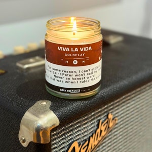 May include: A candle with a brown label that says "Viva La Vida" and "Coldplay". The label also has song lyrics from the song "Viva La Vida" by Coldplay. The candle is on a black surface.
