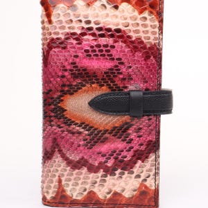 May include: A vertical wallet with a snakeskin pattern in shades of pink, red, and tan. The wallet has a black leather strap closure. The design combines natural textures and vibrant colours, creating a unique accessory.