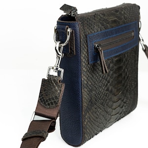 Pode incluir: Bolsa transversal retangular com um padrão de pele de cobra cinza escuro texturizado. A bolsa apresenta um painel lateral de couro azul e um detalhe azul no bolso frontal com zíper. Possui uma alça ajustável marrom e ferragens prateadas.
