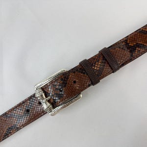 May include: A brown and black snakeskin-patterned belt with a silver-colored buckle. The belt has a dark brown leather keeper and a silver buckle with decorative detailing. The snakeskin pattern features shades of brown, black, and gray.