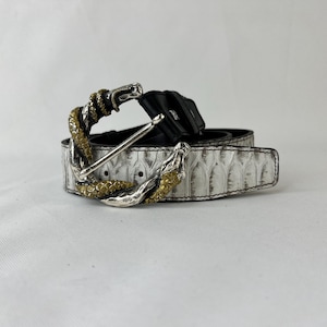 May include: A white leather belt with a snake-shaped buckle. The buckle is silver and gold, with the snake's head and tail wrapping around the belt. The belt has a black interior and a textured, patterned exterior.