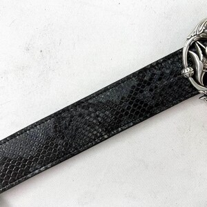 May include: Black belt with a snakeskin pattern. The belt has a silver-colored buckle with an ornate floral design. The belt is made of a textured material and is set against a white background.