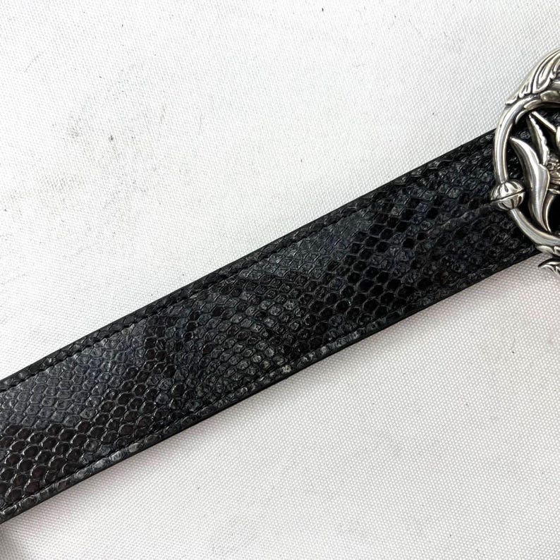 May include: Black belt with a snakeskin pattern. The belt has a silver-colored buckle with an ornate floral design. The belt is made of a textured material and is set against a white background.