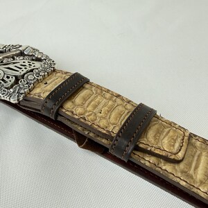 K&ouml;nnte beinhalten: Nahaufnahme eines G&uuml;rtels mit silberner Schnalle und hellbraunem Schlangenmuster. Der G&uuml;rtel hat dunkelbraune Lederakzente und eine dunkelrote Unterseite. Die Schnalle hat ein aufwendiges Design.