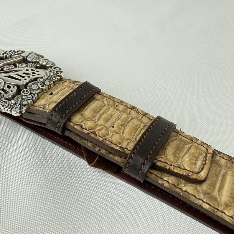 K&ouml;nnte beinhalten: Nahaufnahme eines G&uuml;rtels mit silberner Schnalle und hellbraunem Schlangenmuster. Der G&uuml;rtel hat dunkelbraune Lederakzente und eine dunkelrote Unterseite. Die Schnalle hat ein aufwendiges Design.