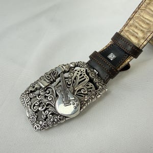 K&ouml;nnte beinhalten: Silberfarbene G&uuml;rtelschnalle mit Gitarrendesign und dem Jahr 1975. Die Schnalle ist an einem braunen Lederg&uuml;rtel mit Schlangenmuster befestigt. Die Schnalle hat ein detailliertes Blumen- und Scroll-Design.