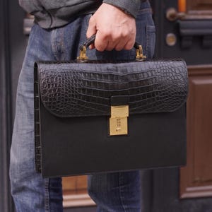 May include: Black leather briefcase with a crocodile-effect flap and gold-coloured hardware. The briefcase has a rectangular shape with a handle and a lock. The briefcase is being held by a person wearing blue jeans and a grey jacket.