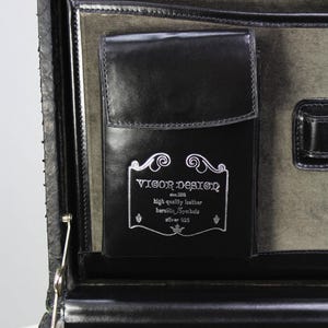 May include: Black leather briefcase with a silver clasp and a pocket with the text "VIGOR DESIGN" and the words "high quality leather". The interior is lined with a gray fabric.