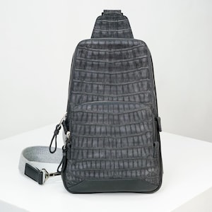 May include: A gray, textured leather sling bag with a crocodile pattern. The bag has a zippered main compartment, a front pocket, and a gray adjustable strap. The bag is approximately 15 inches tall and 8 inches wide.
