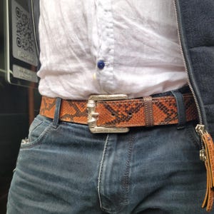 May include: A snakeskin belt with a silver buckle is worn with blue jeans and a white shirt. The belt features a brown and black snakeskin pattern. The silver buckle is rectangular and has a decorative design.