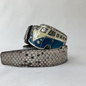 May include: A belt featuring a silver buckle in the shape of a blue and cream-coloured vintage bus. The belt is grey with a snakeskin pattern. The buckle has black detailing.