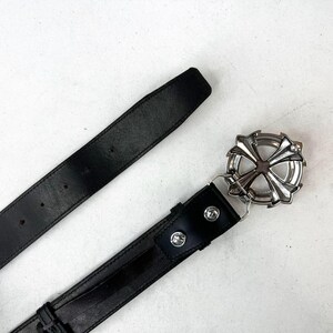 May include: Black leather belt with a silver-coloured, ornate buckle. The buckle has a cross-like design with a circular frame. The belt is shown against a white background.