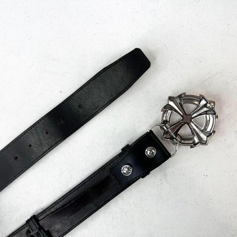May include: Black leather belt with a silver-coloured, ornate buckle. The buckle has a cross-like design with a circular frame. The belt is shown against a white background.