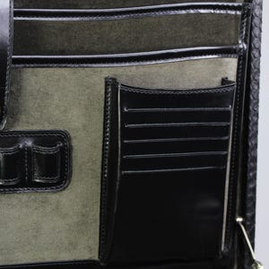 May include: Close-up of the interior of a black leather briefcase. The interior features multiple pockets, including card slots, and a soft, olive-green suede lining. The briefcase has a classic design with a secure closure and is likely used for professional purposes.