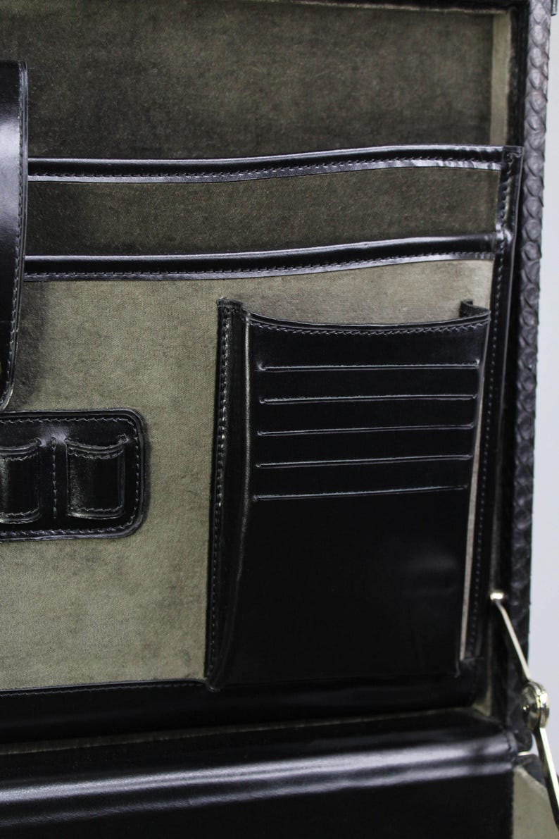 May include: Close-up of the interior of a black leather briefcase. The interior features multiple pockets, including card slots, and a soft, olive-green suede lining. The briefcase has a classic design with a secure closure and is likely used for professional purposes.