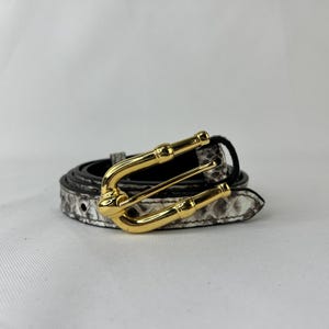 May include: A coiled belt with a snakeskin pattern in shades of gray and white. The belt features a gold-colored buckle with a bamboo-like design. The belt is likely made of leather or a similar material.