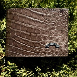 May include: A bi-fold wallet featuring a textured, grey, crocodile-embossed pattern. The front surface displays a small, silver-toned metal emblem shaped like a curved banner with the text "VISOR DESIGN" and "since 1999" engraved on its face.