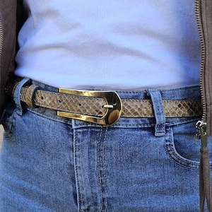 May include: A snakeskin-patterned belt with a gold-coloured buckle is worn over blue jeans. The belt is tan and brown, and the buckle is a shiny gold colour. The jeans are a medium wash denim.