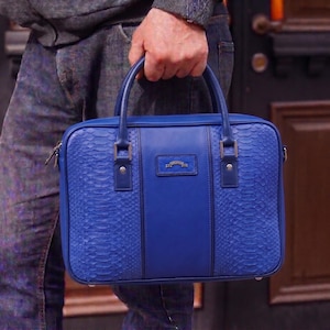 Peut inclure: Une mallette bleu vif avec une texture peau de serpent sur les côtés et un panneau central lisse. Le sac a deux poignées et un petit emblème rectangulaire. La mallette est tenue par une personne.
