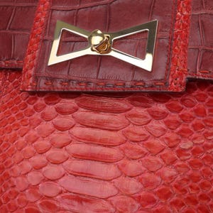 Peut inclure: Gros plan sur une pochette rouge avec un motif texturé en forme de peau de serpent. La pochette est ornée d'un fermoir doré en forme de nœud. Le rabat supérieur est rouge plus foncé avec une texture peau de crocodile.