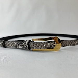 Könnte beinhalten: Ein Gürtel mit Schlangenmuster und einer goldfarbenen rechteckigen Schnalle. Der Gürtel ist hauptsächlich grau und schwarz mit einem dünnen schwarzen Riemen. Die Schnalle hat eine glänzende Goldfarbe.