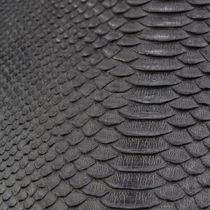 May include: Close-up of a textured, dark gray material with a scale-like pattern. The scales overlap, creating a reptile skin appearance. The surface has a subtle sheen, suggesting a leather or similar material.