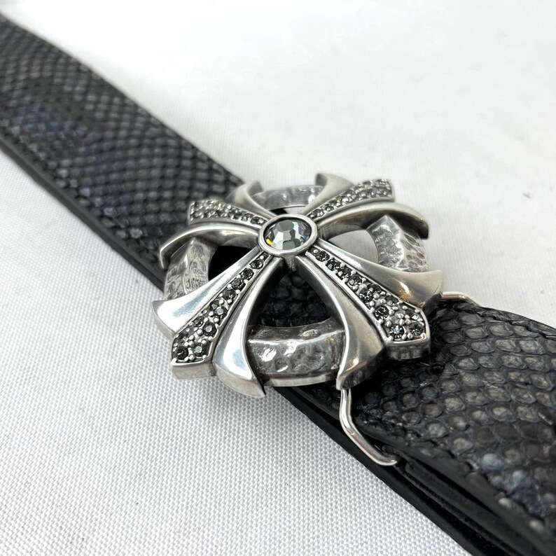 May include: A silver belt buckle featuring a cross design, adorned with small black stones and a central green gem. The buckle is fastened to a black belt with a textured snakeskin pattern.
