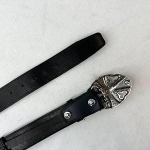 K&ouml;nnte beinhalten: Schwarzer Lederg&uuml;rtel mit silberner Schnalle. Die Schnalle hat ein dekoratives Design mit kreuzf&ouml;rmiger Gestaltung. Der G&uuml;rtel wird vor wei&szlig;em Hintergrund gezeigt.