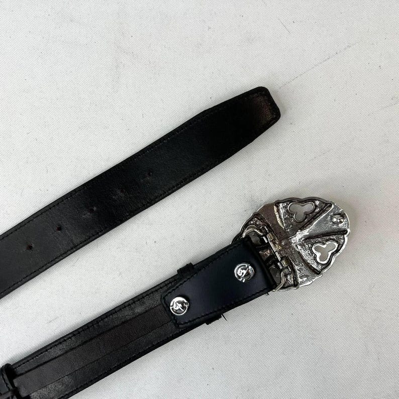 K&ouml;nnte beinhalten: Schwarzer Lederg&uuml;rtel mit silberner Schnalle. Die Schnalle hat ein dekoratives Design mit kreuzf&ouml;rmiger Gestaltung. Der G&uuml;rtel wird vor wei&szlig;em Hintergrund gezeigt.