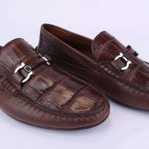 May include: A pair of brown leather loafers with a crocodile-textured vamp and a silver-colored buckle. The shoes have a classic design and are suitable for casual or semi-formal wear.