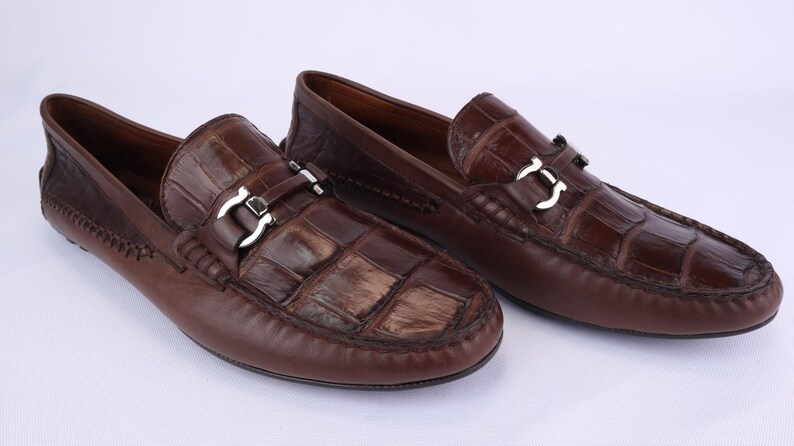 May include: A pair of brown leather loafers with a crocodile-textured vamp and a silver-colored buckle. The shoes have a classic design and are suitable for casual or semi-formal wear.