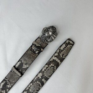 Pode incluir: Um cinto com padr&atilde;o de pele de cobra com uma fivela prateada. A fivela apresenta um design floral detalhado. O cinto &eacute; em tons de cinza e castanho, com um aspeto texturizado. O cinto est&aacute; desabotoado e sobre uma superf&iacute;cie branca.