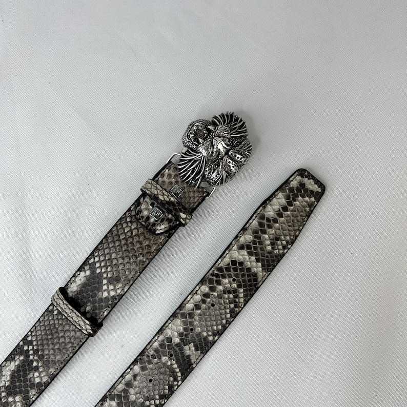 Pode incluir: Um cinto com padr&atilde;o de pele de cobra com uma fivela prateada. A fivela apresenta um design floral detalhado. O cinto &eacute; em tons de cinza e castanho, com um aspeto texturizado. O cinto est&aacute; desabotoado e sobre uma superf&iacute;cie branca.