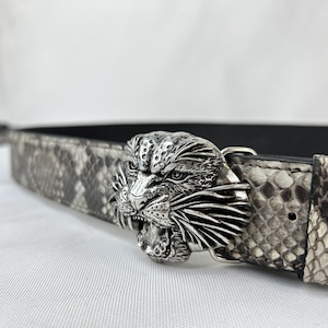 Pode incluir: Uma fivela de cinto de metal prateado em forma de cabe&ccedil;a de jaguar rugindo &eacute; o ponto focal deste cinto. O pr&oacute;prio cinto tem um design de pele de cobra em tons de cinza e preto. Um acess&oacute;rio elegante.