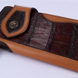 May include: A brown and tan leather wallet with a textured crocodile pattern. The wallet features a decorative skull charm and a flap with a metal emblem. The edges are stitched with tan thread.