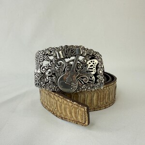 Op de afbeelding: Een zilverkleurige gesp met een gitaarontwerp en de tekst "1973". De riem heeft een lichtbruin slangenpatroon. De gesp is ingewikkeld gedetailleerd met bloemen- en muziekinstrumentmotieven.