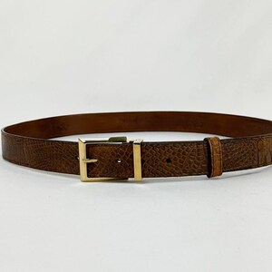 May include: Brown leather belt with a textured, embossed pattern. The belt features a gold-colored rectangular buckle and a matching belt loop. The belt is a classic accessory for trousers or jeans.