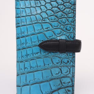 Puede incluir: Una cartera azul turquesa con textura de piel de cocodrilo. La cartera tiene un cierre de correa de cuero negro. La cartera es rectangular y parece estar hecha de cuero.