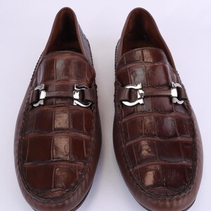 May include: A pair of brown leather loafers with a crocodile pattern. Each shoe features a silver-colored buckle across the vamp. The shoes are displayed on a white background.