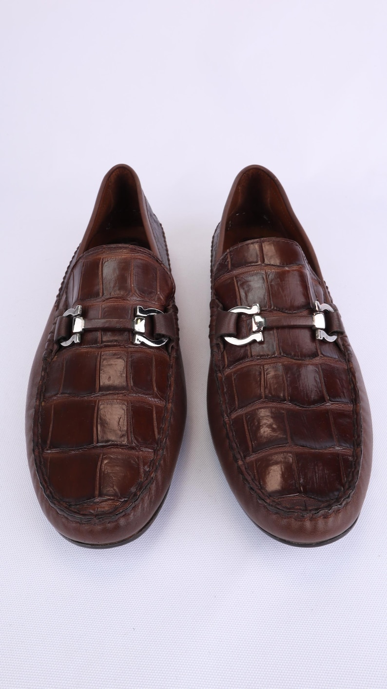 May include: A pair of brown leather loafers with a crocodile pattern. Each shoe features a silver-colored buckle across the vamp. The shoes are displayed on a white background.