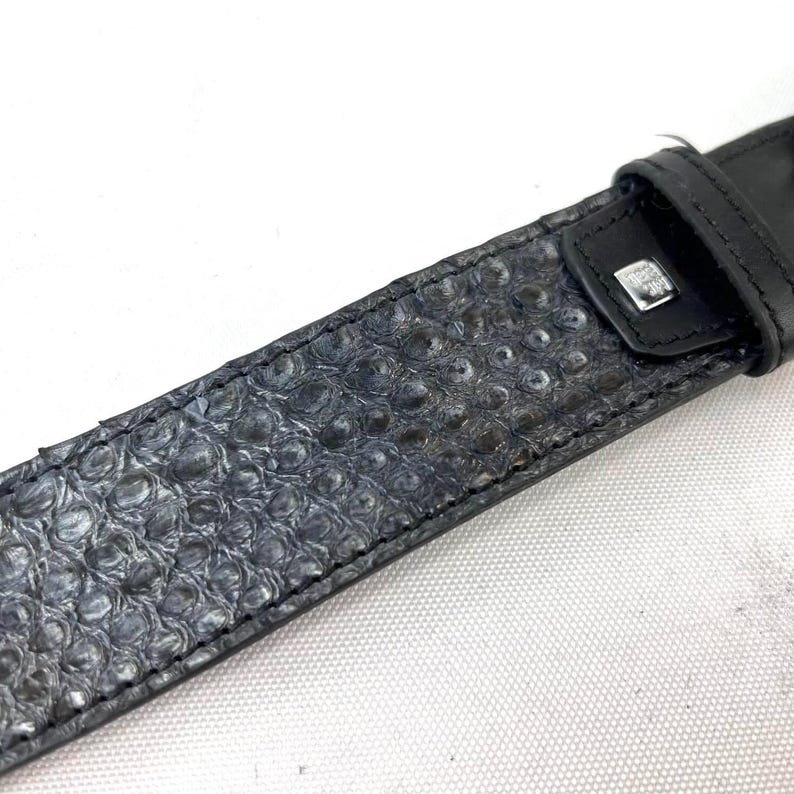 K&ouml;nnte beinhalten: Nahaufnahme eines strukturierten, dunkelgrauen G&uuml;rtels. Der G&uuml;rtel weist ein Muster aus erhabenen, kreisf&ouml;rmigen Erhebungen auf. Das Schnallenende ist aus schwarzem Leder mit einem kleinen, silberfarbenen quadratischen Detail.