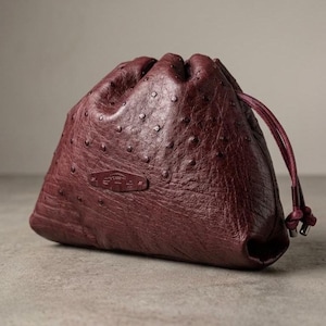 Könnte beinhalten: Ein weinroter Lederbeutel mit Kordelzug, strukturierter Oberfläche und kleinem geprägtem Emblem. Der Beutel hat einen Kordelzugverschluss und ein geknotetes Ende. Die Tasche ist aus weichem Leder gefertigt.