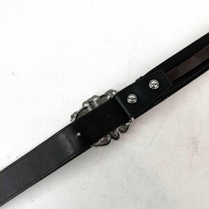 May include: Black leather belt with a decorative silver-tone buckle. The buckle has an ornate, floral design. The belt has a classic, simple design with a single belt loop and two silver-tone rivets near the buckle.