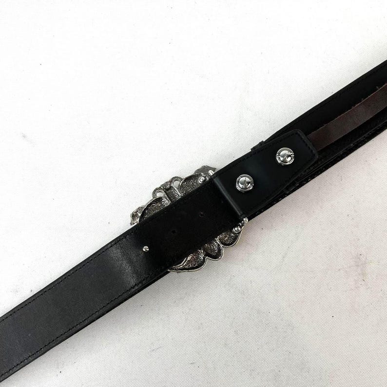 May include: Black leather belt with a decorative silver-tone buckle. The buckle has an ornate, floral design. The belt has a classic, simple design with a single belt loop and two silver-tone rivets near the buckle.