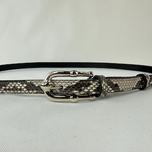 Peut inclure: Une ceinture à motif peau de serpent avec une boucle argentée de style bambou. La ceinture présente un dos en cuir noir et une palette de couleurs neutres de gris, blanc et noir. La boucle est en métal brillant et poli.