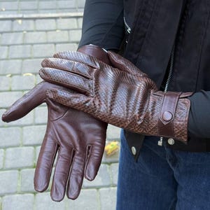 May include: A pair of brown leather gloves with a snakeskin pattern on the top of the hand. The gloves have a smooth leather palm and a strap with a button closure at the wrist. The gloves are a rich, dark brown color.