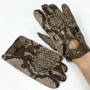 Puede incluir: Un par de guantes de conducción con estampado de piel de serpiente en tonos marrón, gris y beige. Los guantes tienen agujeros para los dedos y una correa de muñeca con cierre de botón.