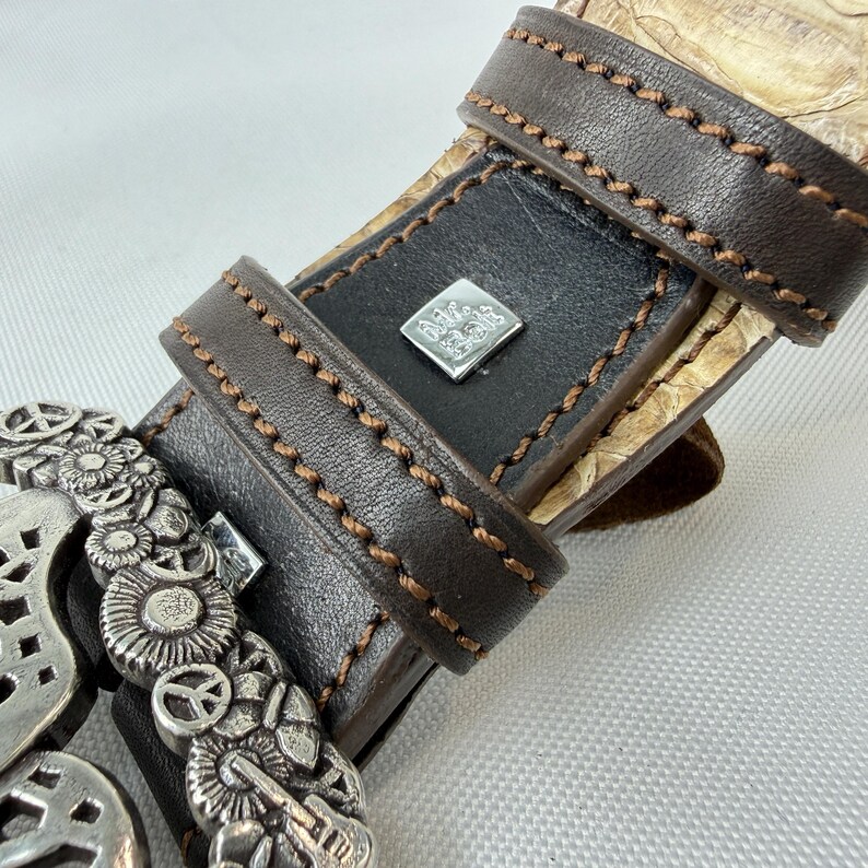 K&ouml;nnte beinhalten: Nahaufnahme eines braunen Lederg&uuml;rtels mit einer dekorativen Silberschnalle. Der G&uuml;rtel hat einen schwarzen Lederabschnitt mit einem quadratischen Silberemblem. Der G&uuml;rtel ist mit braunem Faden gen&auml;ht und hat ein Schlangenmuster.